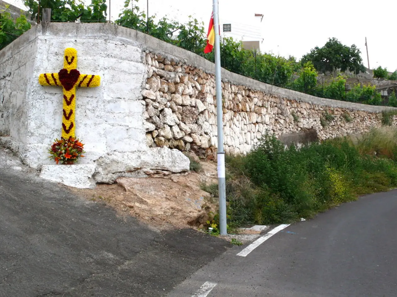 There is a road at the right. There are plants, pole, flag, Christianity sign and flowers. There is...