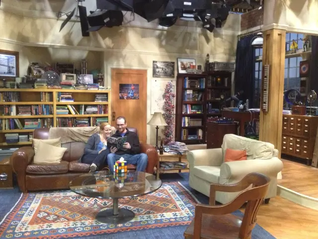 There are sitting in a room. They are sitting in a sofa. He is holding book. She is look at the...