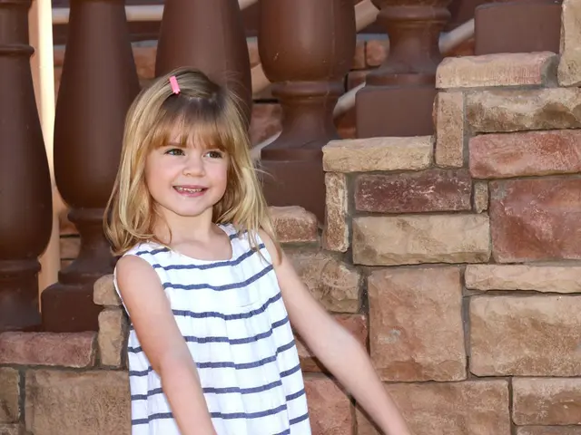 In this image in the front there is a girl standing and smiling. In the background there is a wall...